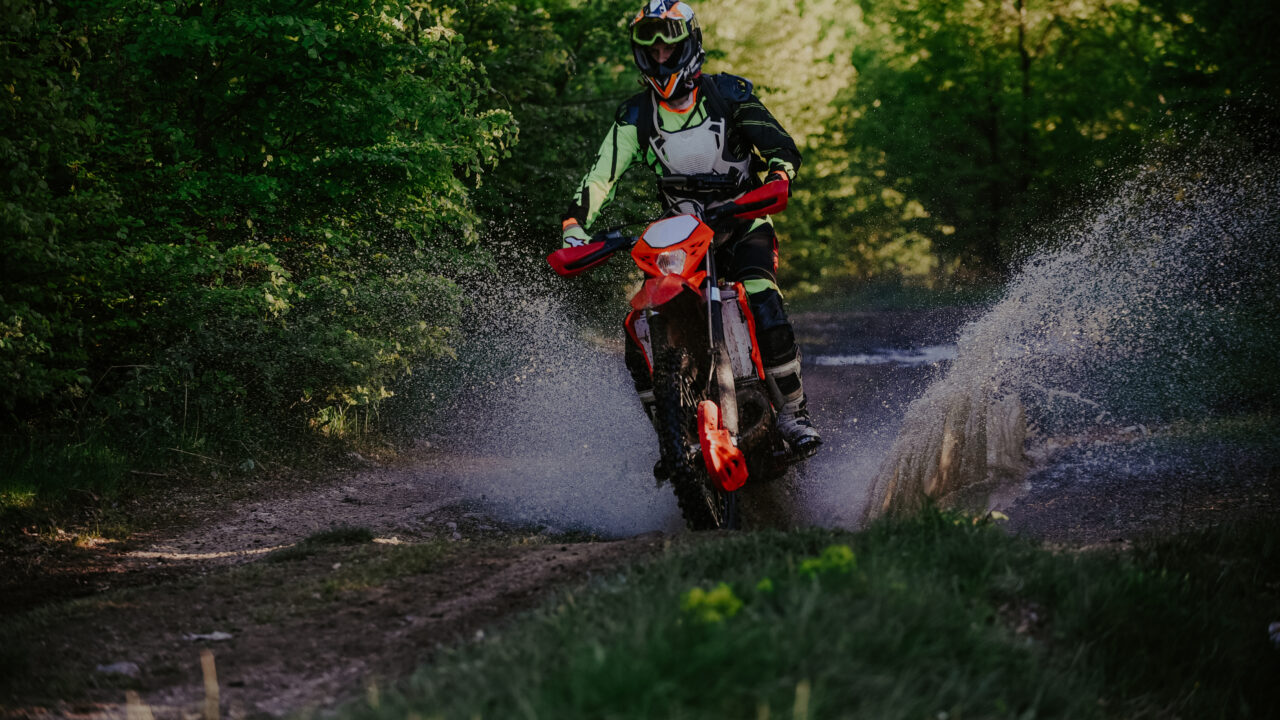 Pilote de motocross en pleine action sur un chemin boueux, moto inclinée dans la terre humide avec projections de boue autour de la roue.
