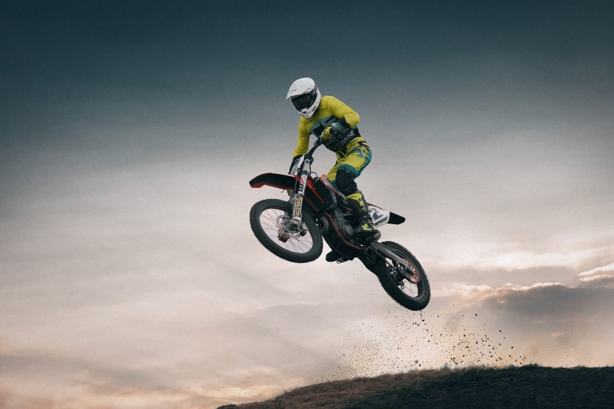 Motocross rider mid-air over a jump in a cross-up, bars turned, dirt thrown off the rear wheel at dusk.