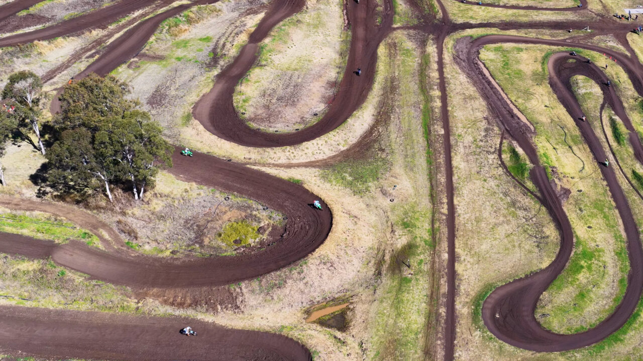 Vue aérienne d'une piste de motocross sinueuse en pleine campagne, avec des pilotes qui roulent sur le tracé en terre.