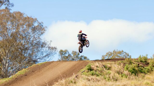 Pilote de motocross en pleine phase d'envol au-dessus d'une bosse de terre ocre, sous un ciel bleu dégagé.