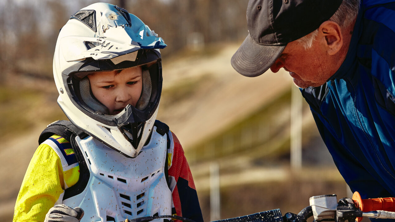 Un moniteur donne des instructions à un jeune pilote de moto avant une compétition junior, tous deux équipés de casques.