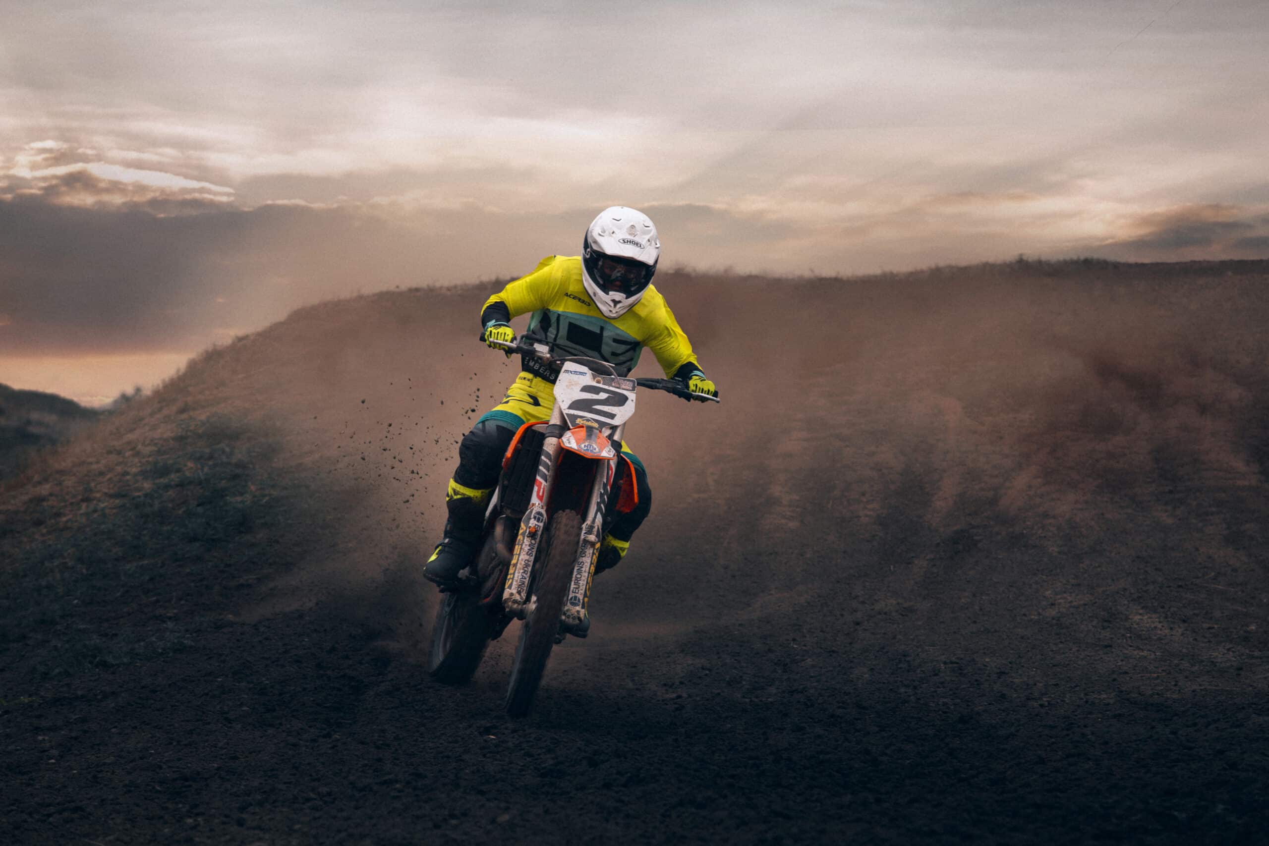 Pilote de motocross en tenue jaune et noire, debout sur les repose-pieds, remontant une pente en terre au crépuscule dans un nuage de poussière.