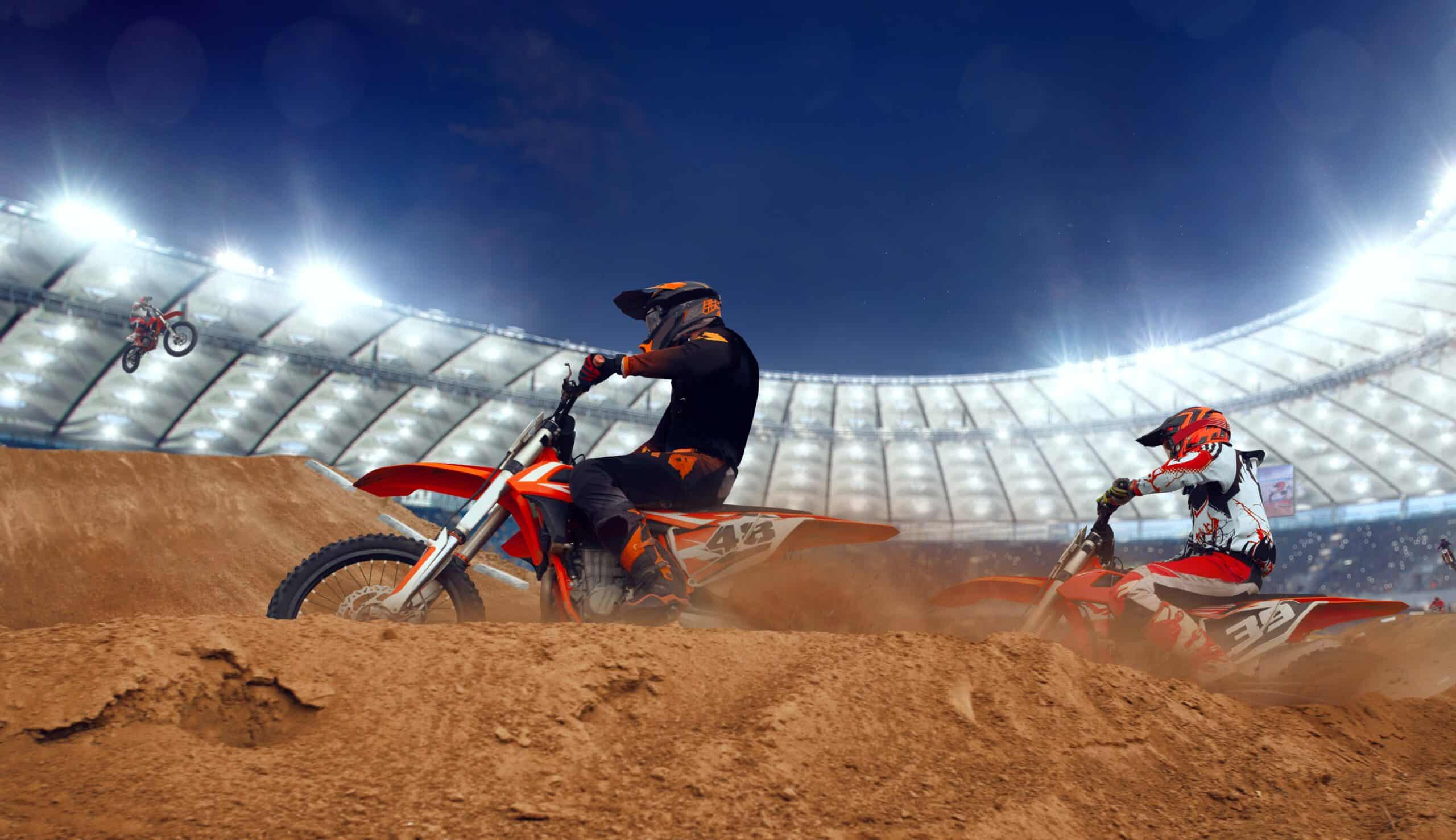 Deux pilotes de motocross en pleine sortie de virage sur une piste en terre, gerbe de poussière soulevée sous un éclairage de stade.