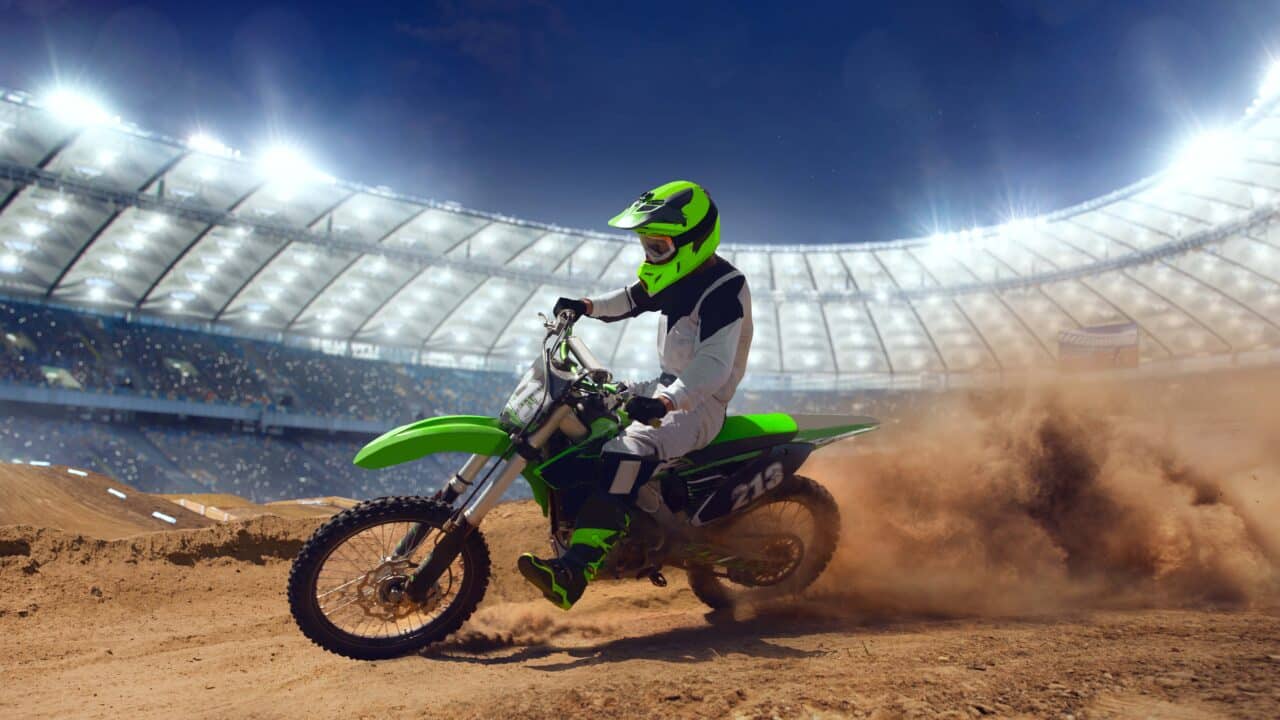 Pilote de motocross en combinaison verte et noire, moto inclinée en sortie de virage sur une piste en terre, gerbe de poussière soulevée sous un éclairage de stade.